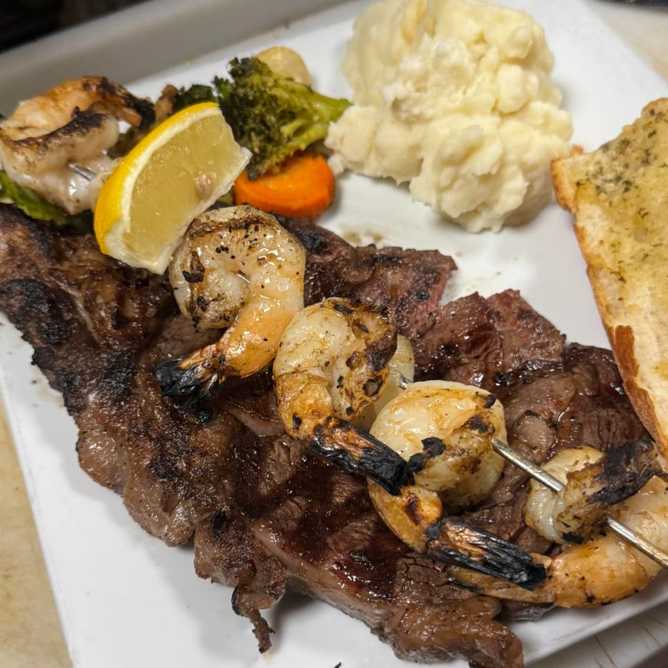 Grilled steak and shrimp skewers with mashed potatoes, roasted vegetables, and garlic bread.