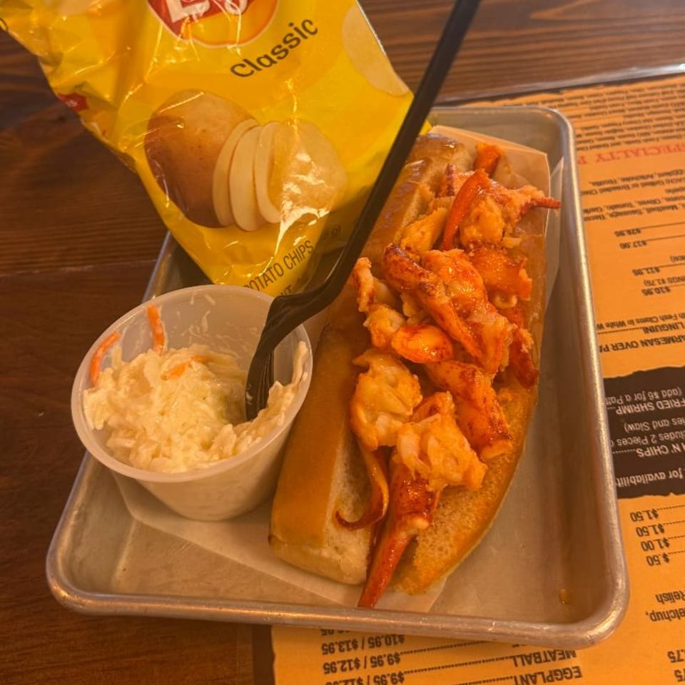 A lobster roll with coleslaw and a bag of Lays potato chips on a metal tray.