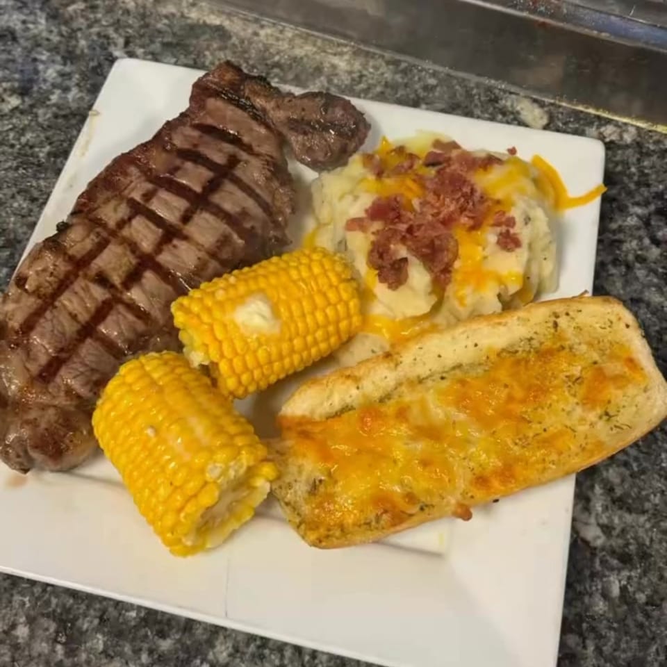 Grilled steak, two corn on the cob, mashed potatoes with bacon, and cheesy garlic bread on a white plate.