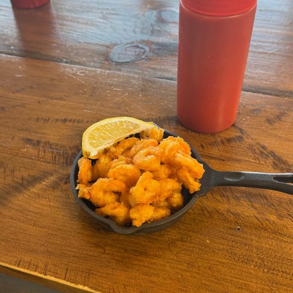 A cast iron skillet of crispy fried shrimp with a lemon wedge, next to a red bottle.