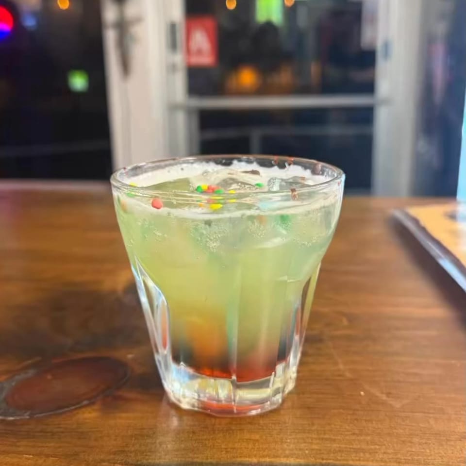A vibrant green cocktail with colorful sprinkles and a red layer at the bottom, on a wooden table.