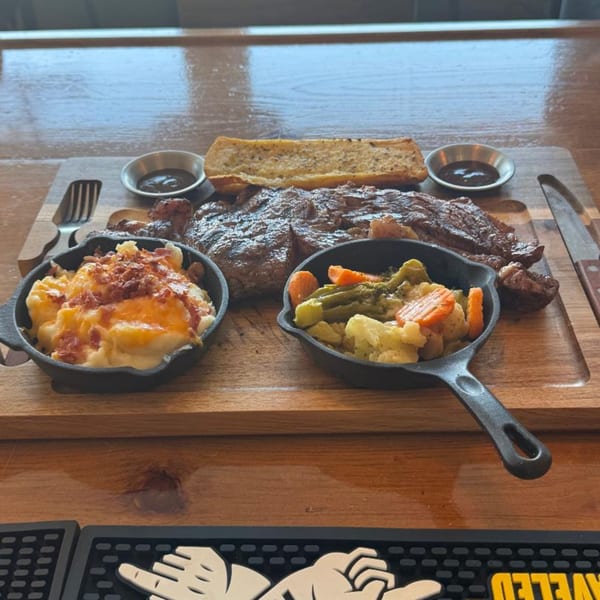 Grilled steak, mac and cheese, and mixed vegetables served on a wooden board with garlic bread.